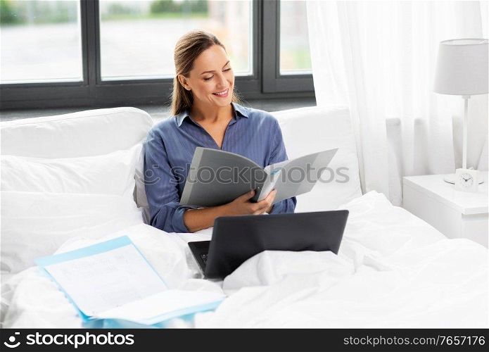 remote job, people and business concept - young woman in glasses with laptop computer and papers in bed working at home. young woman with laptop and papers in bed at home
