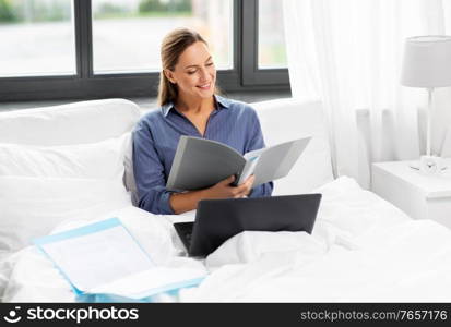 remote job, people and business concept - young woman in glasses with laptop computer and papers in bed working at home. young woman with laptop and papers in bed at home