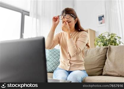 remote job, stress and people concept - tired young asian woman in glasses with laptop computer working and rubbing her nose bridge at home office. tired woman with laptop working at home office