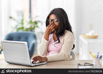 remote job, technology and e-learning concept - tired young african american woman in glasses with laptop computer working at home office and rubbing nose bridge. tired woman with laptop working at home office