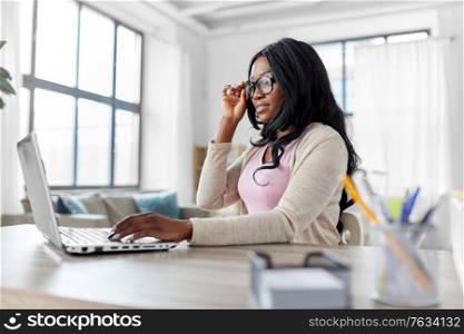 remote job, technology and people concept - african american young woman in glasses with laptop computer working at home office. woman with laptop working at home office