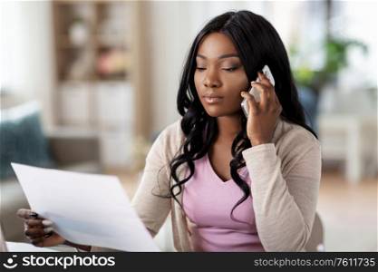 remote job, technology and people concept - african american young woman with laptop computer and papers working at home office and calling on smartphone. woman with papers calling on phone at home office
