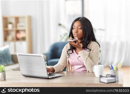 remote job, technology and people concept - african american young woman with laptop computer working at home office and using voice command recorder on smartphone. woman with laptop calling on phone at home office