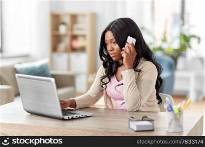 remote job, technology and people concept - african american young woman with laptop computer working at home office and calling on smartphone. woman with laptop calling on phone at home office