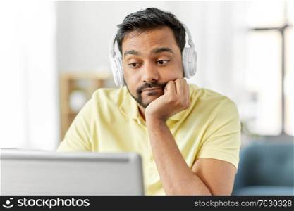 remote job, technology and people concept - bored young indian man in headphones with laptop computer working at home office. bored man in headphones with laptop works at home