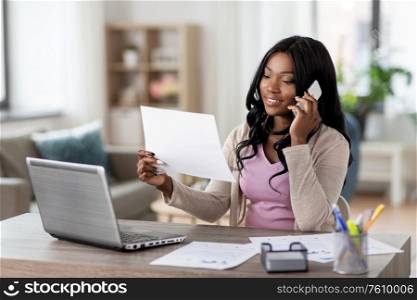 remote job, technology and people concept - happy smiling african american young woman with laptop computer and papers working at home office and calling on smartphone. woman with papers calling on phone at home office