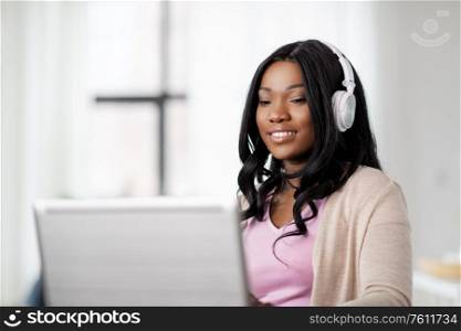 remote job, technology and people concept - happy smiling african american young woman in headphones with laptop computer working at home office. woman in headphones with laptop working at home