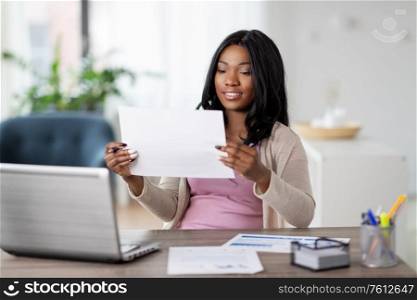 remote job, technology and people concept - happy smiling african american young woman with laptop computer and papers working at home office. happy woman with laptop and papers at home office