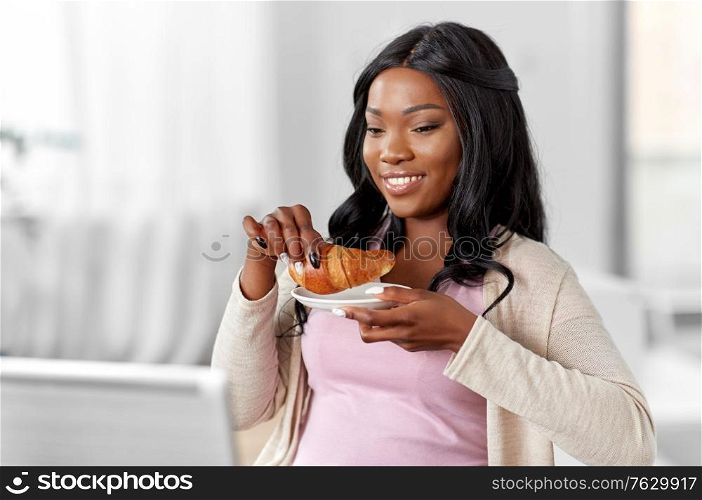 remote job, technology and people concept - happy smiling african american young woman with laptop computer eating croissant at home office. woman with laptop eating croissant at home office
