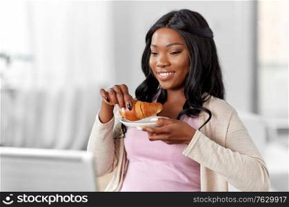 remote job, technology and people concept - happy smiling african american young woman with laptop computer eating croissant at home office. woman with laptop eating croissant at home office