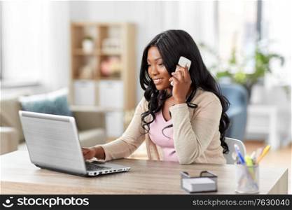 remote job, technology and people concept - happy smiling african american young woman with laptop computer working at home office and calling on smartphone. woman with laptop calling on phone at home office