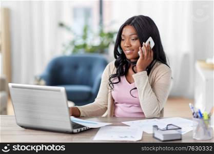 remote job, technology and people concept - happy smiling african american young woman with laptop computer and papers working at home office and calling on smartphone. woman with laptop calling on phone at home office
