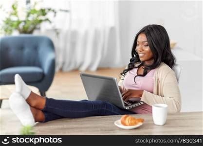 remote job, technology and people concept - happy smiling african american young woman with laptop computer working at home office and resting her feet on table. happy woman with laptop working at home office
