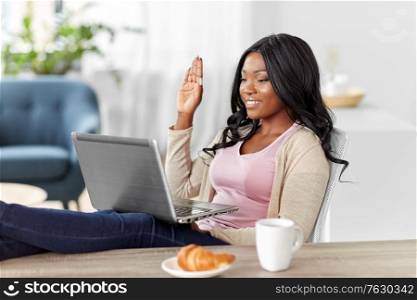 remote job, technology and people concept - happy smiling african american young woman with laptop computer having video call at home office and resting her feet on table. happy woman with laptop working at home office