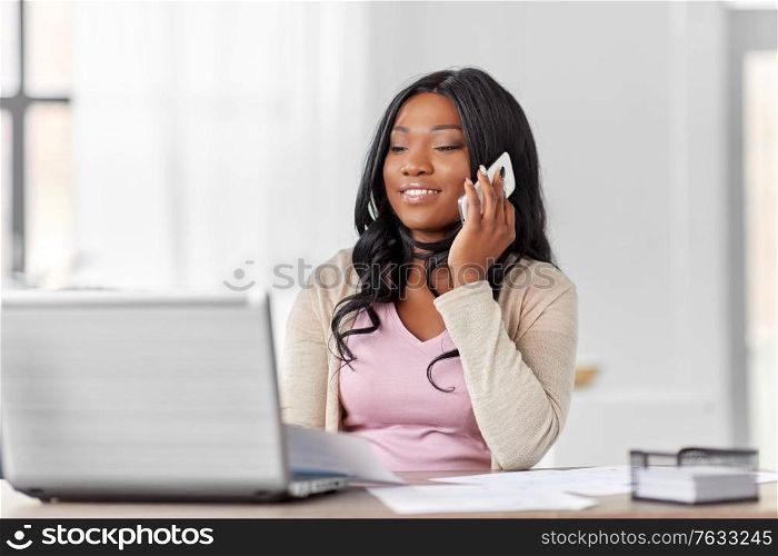 remote job, technology and people concept - happy smiling african american young woman with laptop computer and papers working at home office and calling on smartphone. woman with laptop calling on phone at home office