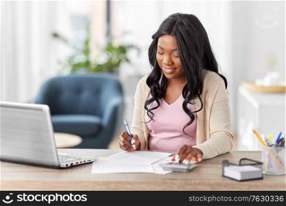remote job, technology and people concept - happy smiling young african american woman with calculator and papers working at home office. woman with calculator and papers working at home