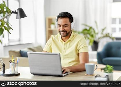 remote job, technology and people concept - happy smiling young indian man in headphones with laptop computer working at home office. man in headphones with laptop working at home