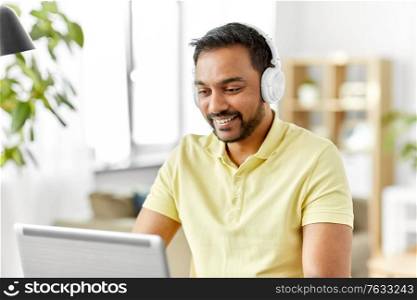 remote job, technology and people concept - happy smiling young indian man in headphones with laptop computer working at home office. man in headphones with laptop working at home