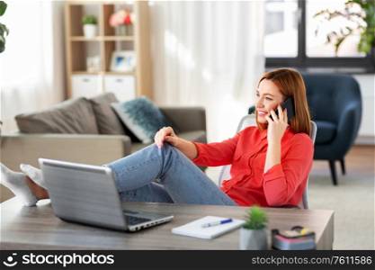 remote job, technology and people concept - happy smiling young woman calling on smartphone with laptop computer at home office resting feet on table. smiling woman calling on smartphone at home office