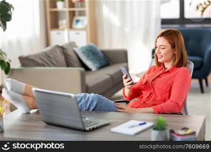 remote job, technology and people concept - happy smiling young woman with smartphone and laptop computer at home office resting feet on table. woman with smartphone and laptop at home office