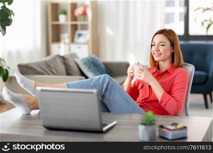 remote job, technology and people concept - happy smiling young woman with laptop computer and coffee having video call at home office with feet on table. woman with laptop drinking coffee at home office