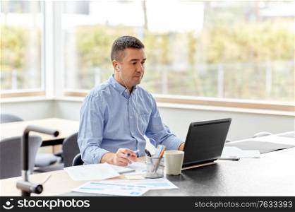 remote job, technology and people concept - middle-aged man in earphones with laptop computer and papers working at home office. man in earphones with laptop working at home