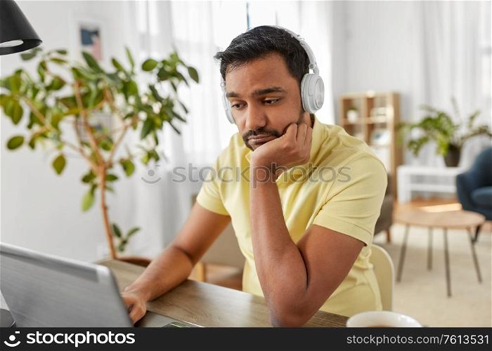 remote job, technology and people concept - sad or bored young indian man in headphones with laptop computer working at home office. man in headphones with laptop working at home