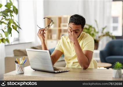 remote job, technology and people concept - tired young indian man in glasses with laptop computer working and rubbing his nose bridge at home office. tired man with laptop working at home office