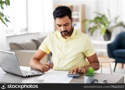 remote job, technology and people concept - young indian man with calculator and papers working at home office. man with calculator and papers working at home