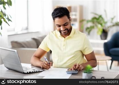 remote job, technology and people concept - young indian man with calculator and papers working at home office. man with calculator and papers working at home