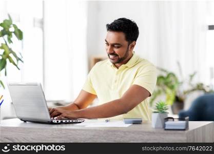 remote job, technology and people concept - young indian man with laptop, calculator and papers working at home office. man with laptop and papers working at home