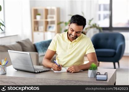 remote job, technology and people concept - young indian man with notebook and laptop computer at home office. indian man with notebook and laptop at home office