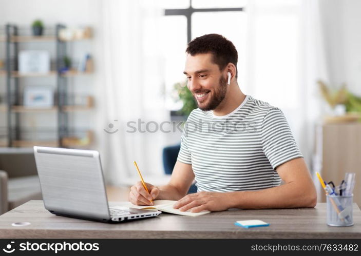 remote job, technology and people concept - young man with earphones, notebook and laptop computer working at home office. man with notebook, earphones and laptop at home
