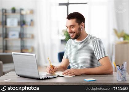 remote job, technology and people concept - young man with earphones, notebook and laptop computer working at home office. man with notebook, earphones and laptop at home