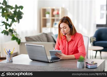 remote job, technology and people concept - young woman with headset and laptop computer having video conference and taking notes to notebook at home office. woman with headset and laptop working at home