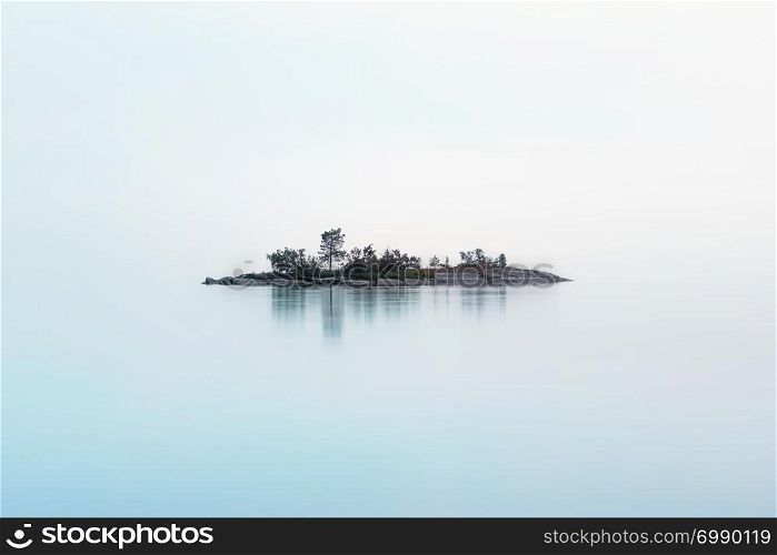 Remote rocky island with coniferous trees as if hanging in the misty air over the milky surface of the lake in a summer night. Atmospheric cool northern background with copy space. White Nights season, Lake Onega, Karelia, Russia.