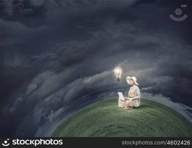Remote work. Woman in dress and hat sitting on green planet and working on laptop