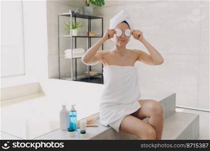 Removing makeup. Funny young woman wrapped in towels fooling, covering eyes with cotton pads during moisturizing or cleansing her face skin from make up, sitting on bathtub in bathroom. . Removing makeup. Girl wrapped in towels cover eyes with cotton pads while cleansing skin in bathroom