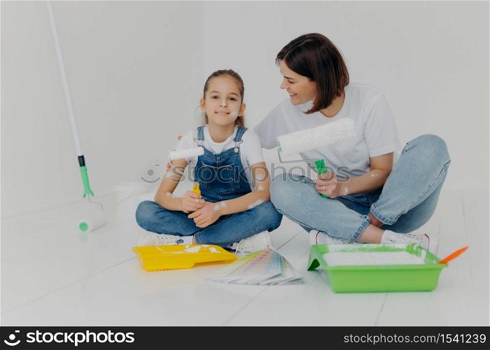 Renovating and remodeling house. Mother and daughter sit together on floor, have rest after painting walls in room, use paint rollers, surrounded with trays and color palette, enjoy teamwork.