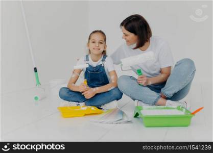 Renovating and remodeling house. Mother and daughter sit together on floor, have rest after painting walls in room, use paint rollers, surrounded with trays and color palette, enjoy teamwork.