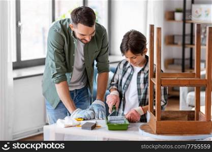 renovation, diy and home improvement concept - father and son in gloves with paint roller painting old wooden table in grey color at home. father and son painting old table in grey color