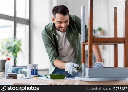 renovation, diy and home improvement concept - happy smiling man in gloves with paint roller painting old wooden table in grey color. man painting old table in grey color at home
