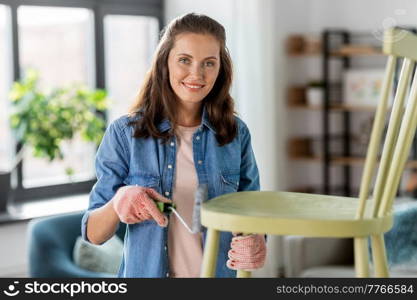 renovation, diy and home improvement concept - happy smiling woman in gloves with paint roller painting old wooden chair in grey color. woman painting old chair in grey color at home