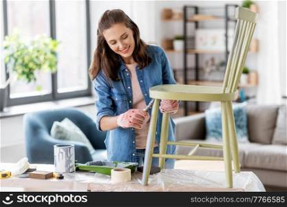 renovation, diy and home improvement concept - happy smiling woman in gloves with paint brush painting old wooden chair in grey color. woman painting old chair in grey color at home
