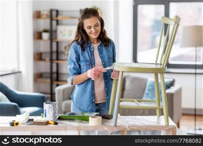renovation, diy and home improvement concept - happy smiling woman in gloves with paint brush painting old wooden chair in grey color. woman painting old chair in grey color at home