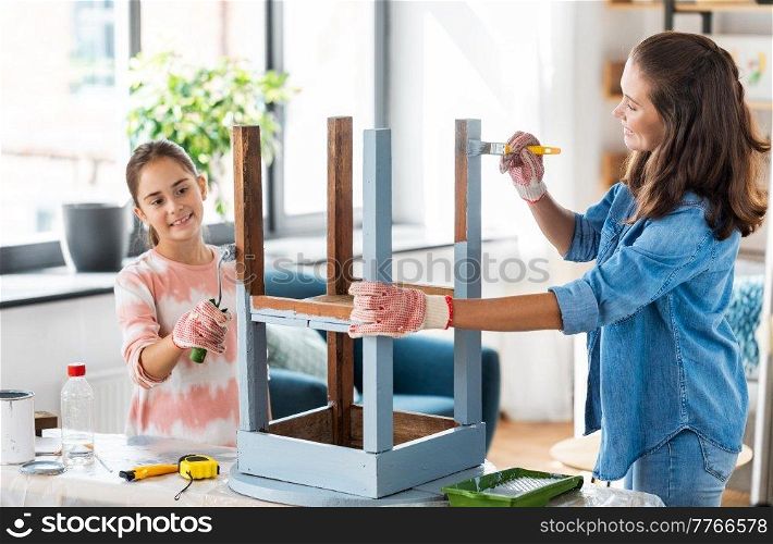 renovation, diy and home improvement concept - mother and daughter in gloves with paint roller and brush painting old wooden table in grey color at home. mother and daughter painting old table at home