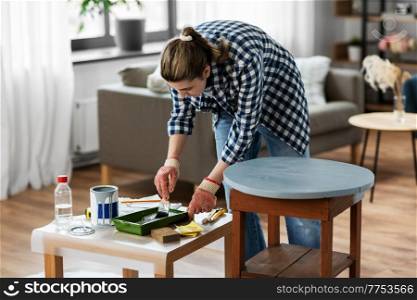 renovation, diy and home improvement concept - woman in gloves with paint brush painting old wooden table to grey color. woman painting old wooden table with grey color