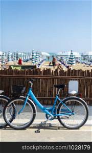 Rental bikes on the beach. Blue bicycles on the street. Many beach umbrellas.
