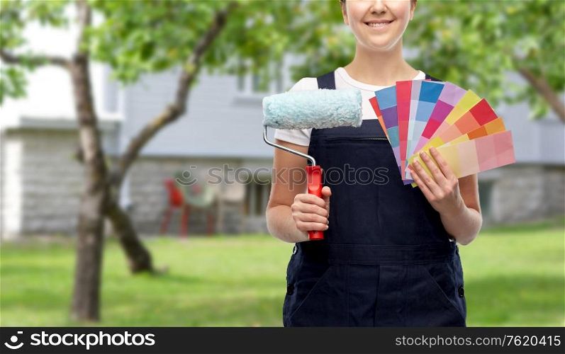 repair, construction and building concept - close up of happy smiling female painter or builder with paint roller and color charts over living house yard with garden background. close up of painter with roller and color charts