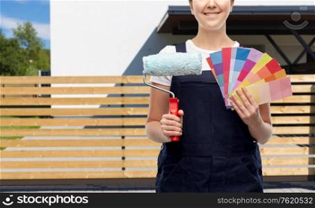 repair, construction and building concept - close up of happy smiling female painter or builder with paint roller and color charts over living house background. close up of painter with roller and color charts